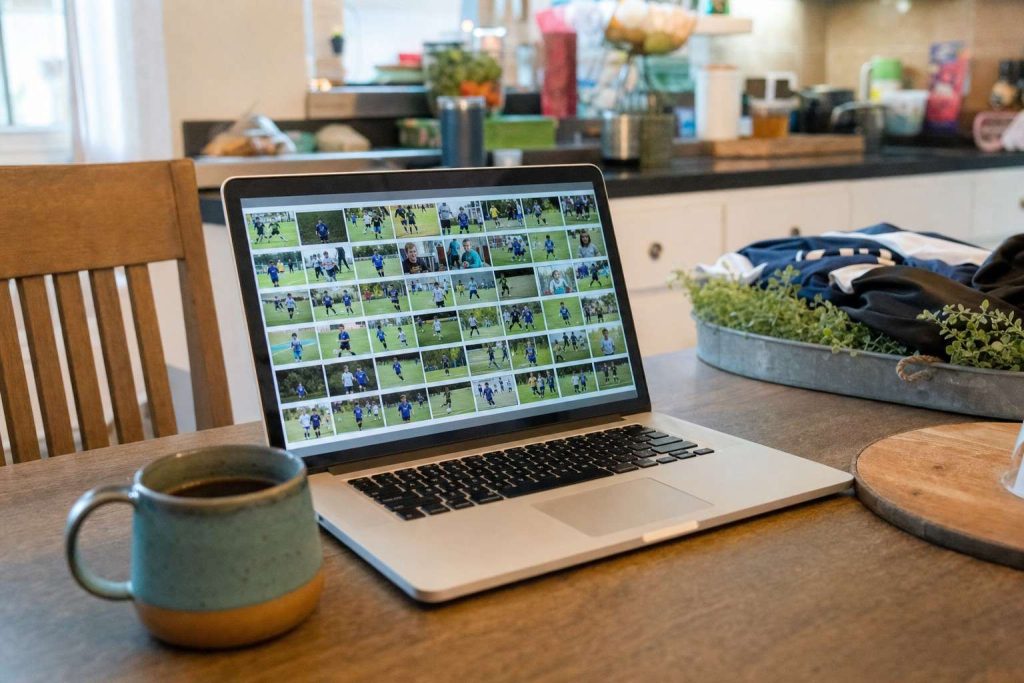 A laptop on a kitchen table showing a grid of small youth sports photo thumbnails